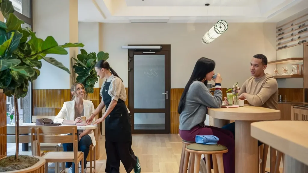 People sit at tables in a modern cafe, some talking and drinking, while a server walks by. The space is bright with wooden decor and green plants.